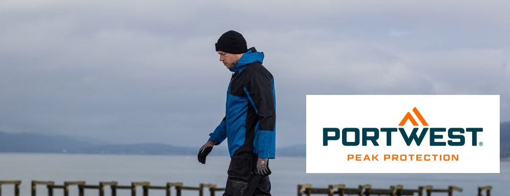 Imagen de un hombre con ropa de trabajo azul y negra y una gorra negra en la cabeza. Está caminando por un sendero de cemento a la orilla de un lago, el cielo detrás de él es gris.