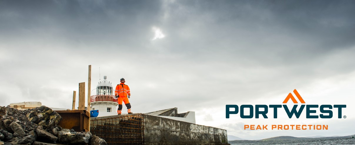 Un trabajador vestido con ropa naranja de alta visibilidad se encuentra en un sitio de construcción rocoso junto a un faro, con el logotipo de Portwest y el lema "Peak Protection" en la imagen.