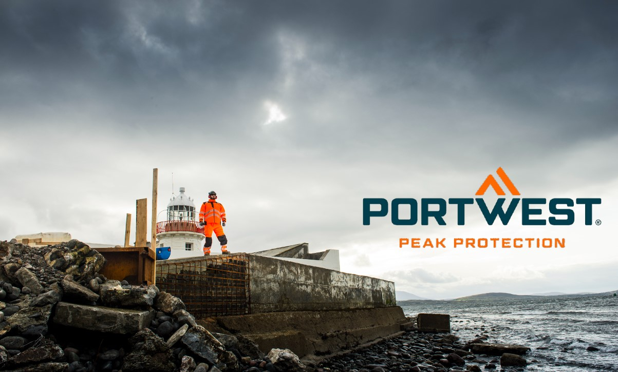 Un trabajador vestido con ropa naranja de alta visibilidad se encuentra en un sitio de construcción rocoso junto a un faro, con el logotipo de Portwest y el lema "Peak Protection" en la imagen. Existe un enlace que conduce a nuestra ropa de trabajo multinorma.