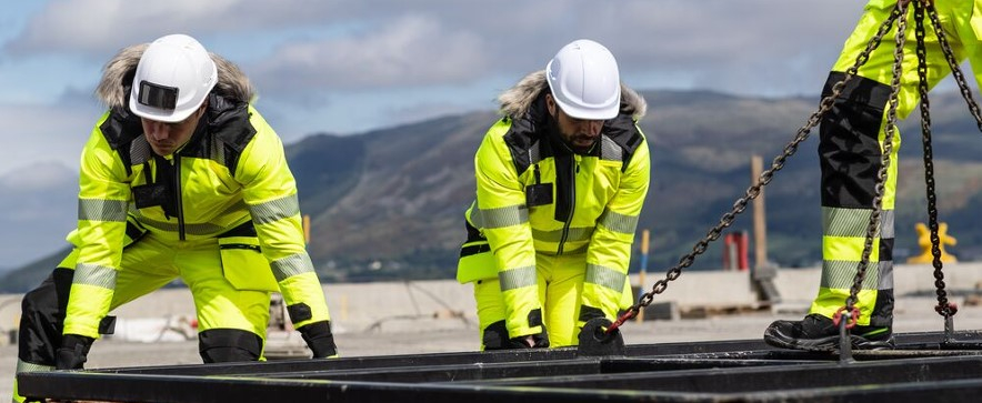 Tres trabajadores con ropa de invierno amarilla y cascos blancos frente a un fondo montañoso con un cielo azul ligeramente nublado.