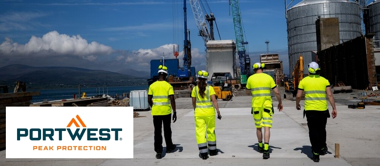 Cuatro trabajadores vestidos con ropa amarilla de alta visibilidad frente a un fondo con silos y grúas. Al fondo se puede ver una cadena montañosa y un cielo azul con nubes blancas dispersas. En el borde inferior izquierdo de la imagen se encuentra el logo de Portwest en azul y naranja sobre un fondo blanco.