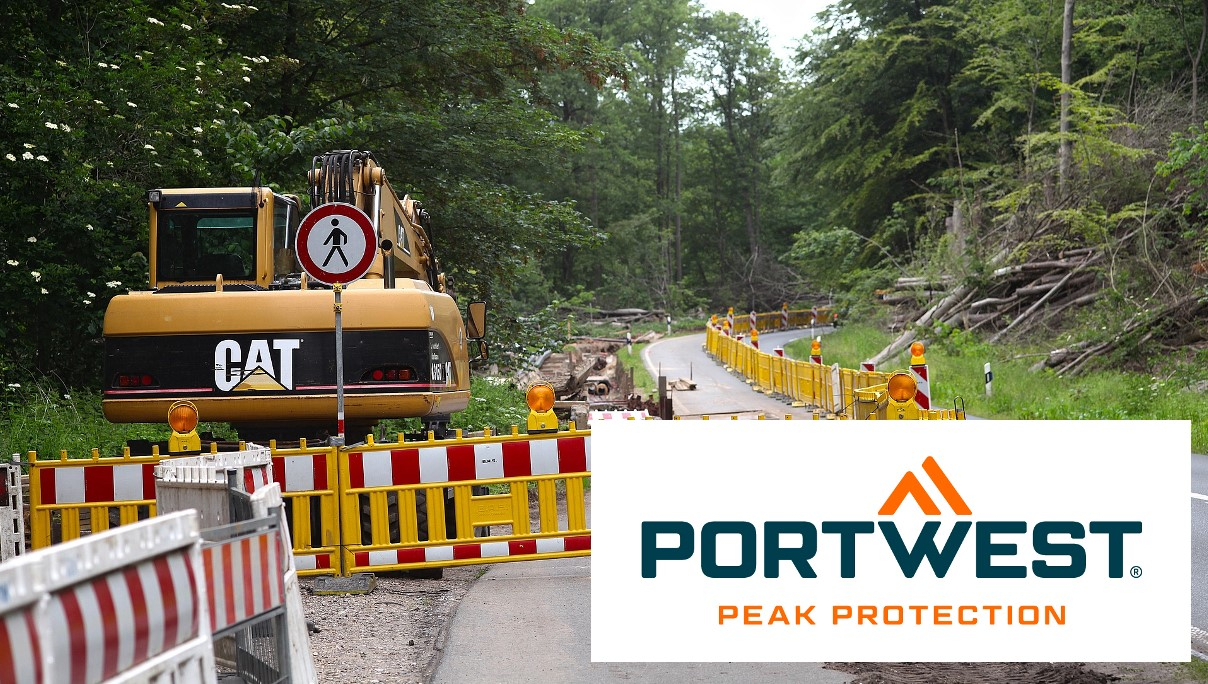 Sitio de construcción en una carretera en el bosque. Una barrera amarilla delimita el sitio de construcción. En la esquina inferior derecha de la imagen se encuentra el logo de Portwest en azul y naranja sobre un fondo blanco. Existe un enlace que le lleva a nuestro departamento de ropa de protección para la construcción de carreteras.