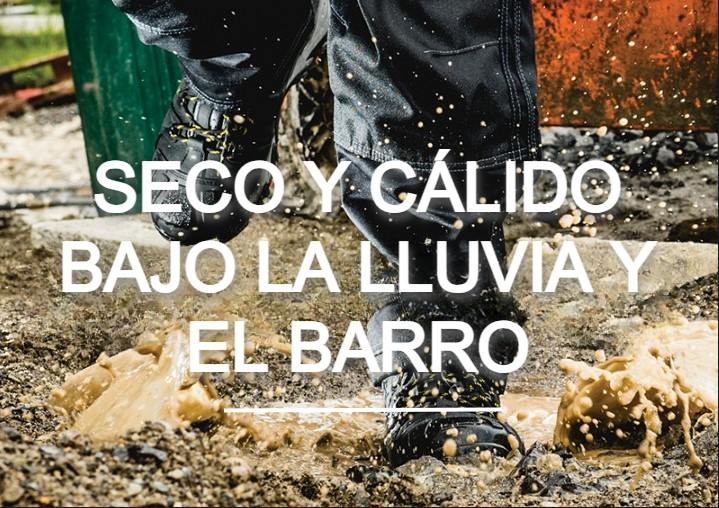 Trabajador con pantalones de lluvia y botas caminando hacia un charco. Salpica barro y al fondo se ve una obra en construcción. Hay un enlace a nuestros zapatos impermeables.