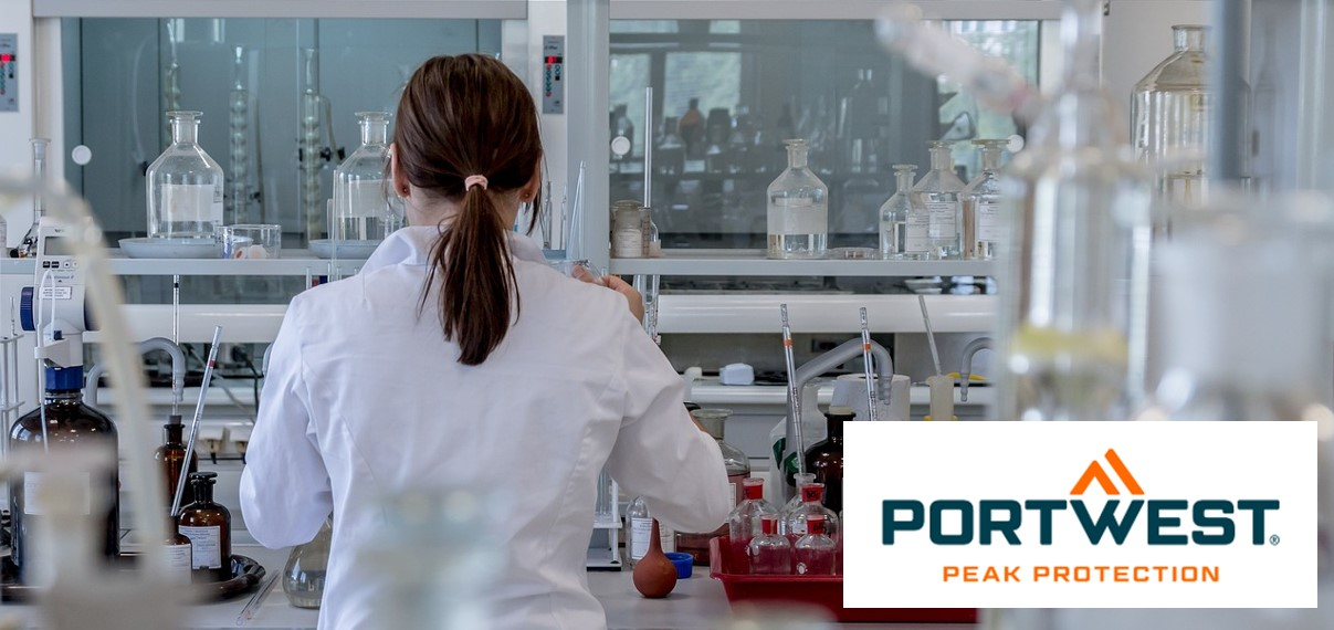 Una mujer con cabello castaño oscuro recogido hacia atrás viste una bata de laboratorio blanca y trabaja en un laboratorio moderno con varios equipos de laboratorio y botellas de productos químicos. En la parte inferior derecha de la imagen se encuentra el logotipo “Portwest Peak Protection”.