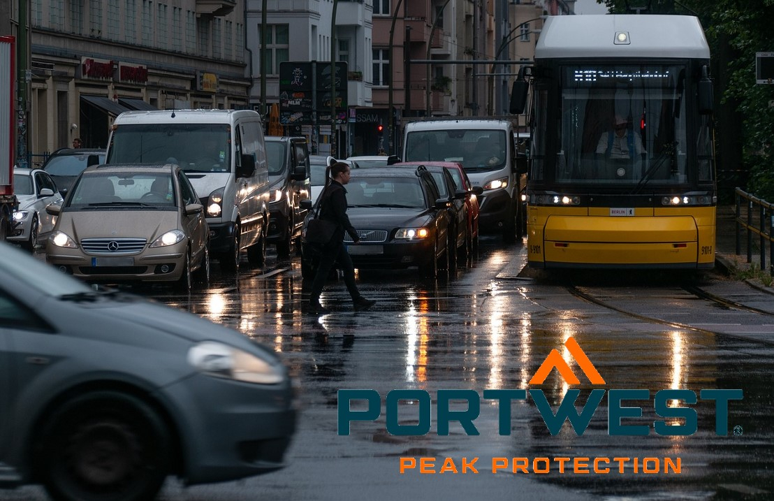 Escena callejera bajo la lluvia. Numerosos coches y autobuses están aparcados en un cruce con las luces encendidas. El logotipo de la empresa Portwest en naranja y azul se encuentra en la esquina inferior derecha de la imagen. Hay un enlace que lleva a nuestros chubasqueros.