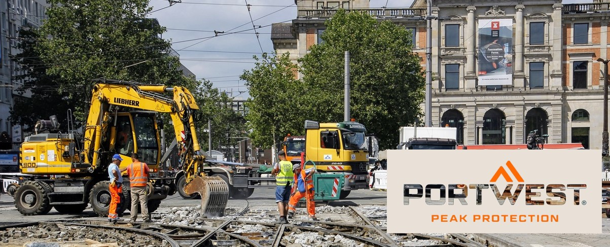 Trabajadores de la construcción con ropa de seguridad naranja trabajan en una obra de construcción en la ciudad. Al fondo se ven excavadoras, camiones y un edificio histórico. El logotipo de Portwest y el lema "Peak Protection" se muestran en la parte inferior de la imagen.