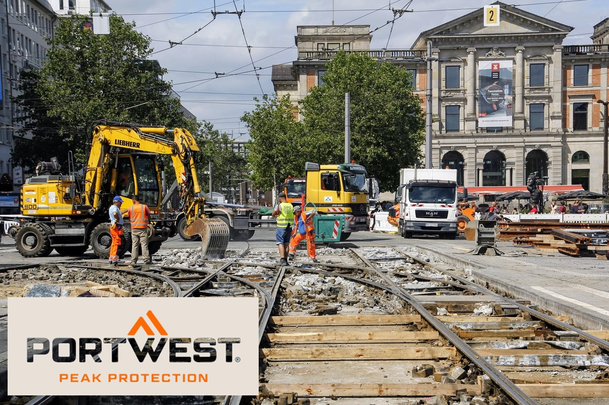 Trabajadores de la construcción con ropa de seguridad naranja trabajan en una obra de construcción en la ciudad. Al fondo se ven excavadoras, camiones y un edificio histórico. El logotipo de Portwest y el lema "Peak Protection" se muestran en la parte inferior de la imagen. Hay un enlace que conduce a la página del artículo sobre el gorro de enfriamiento.